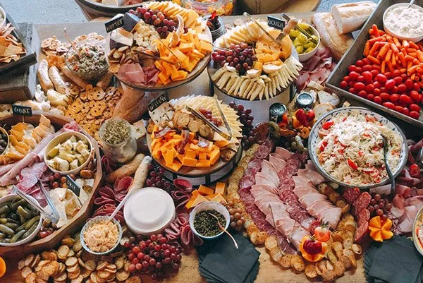 A table filled with a variety of party foods including cheese platters, cured meats, fresh fruit, cherry tomatoes, pickles, crackers, and dips.