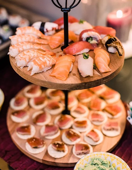Two-tiered serving tray with assorted sushi, including nigiri and sashimi, on a wooden platter with a blurred background, candle, and small bowl of wasabi.