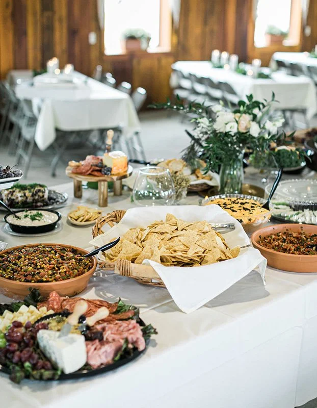 A buffet table with chips, dips, and salads in a rustic wooden venue with white tablecloths and floral centerpieces.