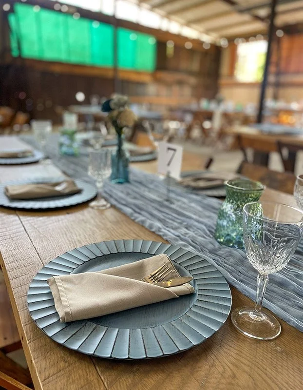 A wedding or event reception table setup with a plate, napkin, fork, and knife, decorated with a table runner, flowers, glassware, and a table number card.