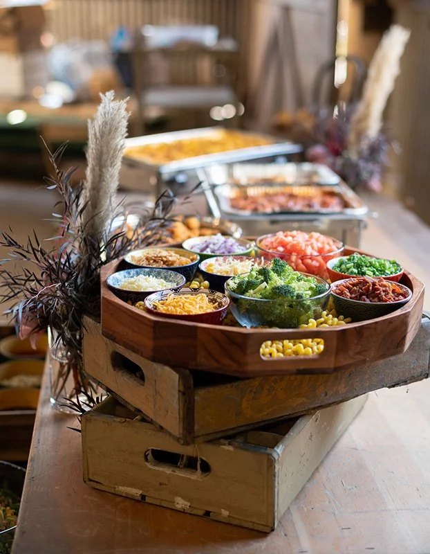 A wooden serving tray filled with various fresh vegetables and toppings, including broccoli, chopped tomatoes, and shredded cheese, on a rustic table. In the background, there are chafing dishes with hot foods in a cozy, homey setting.