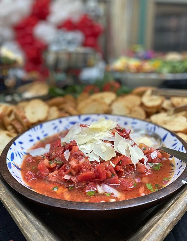 A bowl of gazpacho topped with shaved cheese, surrounded by bread slices, on a wooden tray at a food market or stall.