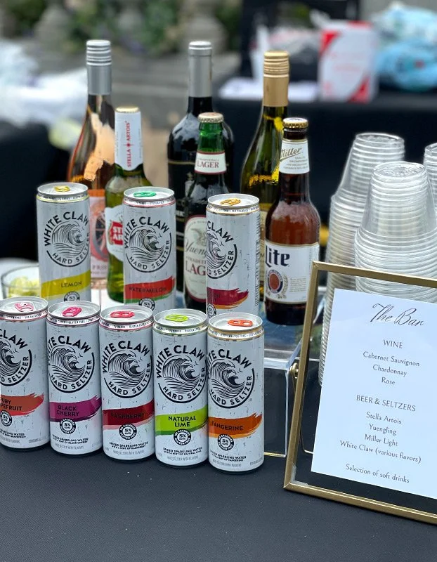 Assorted canned White Claw hard seltzers, small bottles of beer and wine, plastic cups, and a menu display on a table at a bar or outdoor event.