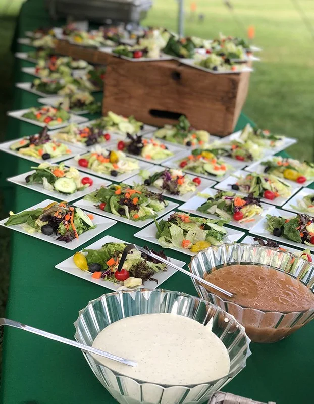 A table covered with small plates of salad and two bowls of dressing at an outdoor event.