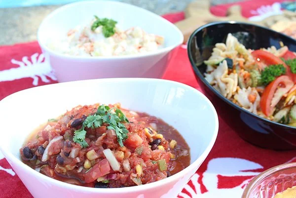 Three bowls of food on a red tablecloth with the background showing a wooden surface. The front bowl contains chili with beans, corn, and tomatoes, garnished with cilantro. The back bowls contain potato salad and a mixed vegetable salad.