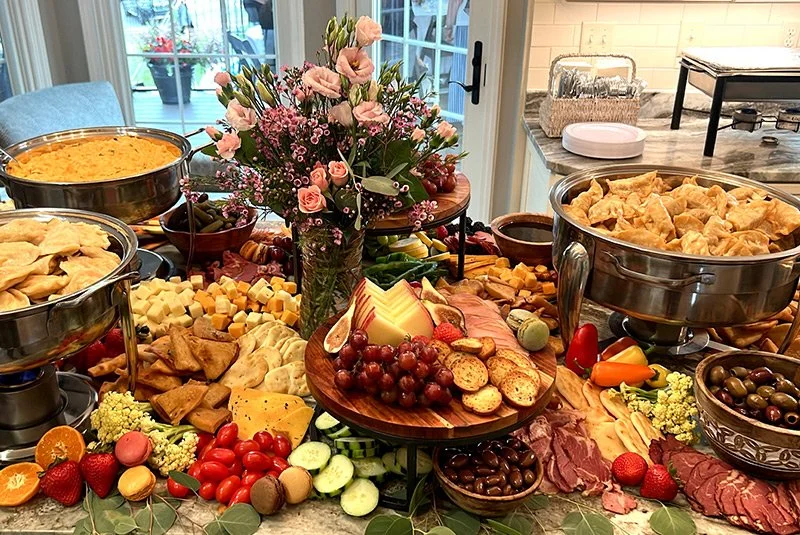 A table set with various food items including cheeses, grapes, cherry tomatoes, sliced cucumbers, crackers, bread, cherry peppers, olives, and bowls of chips, with a centerpiece of pink and purple flowers, in a bright room with a sliding glass door i
