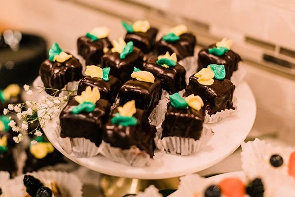 Chocolate petit fours topped with yellow and green decorations on a white cake stand.