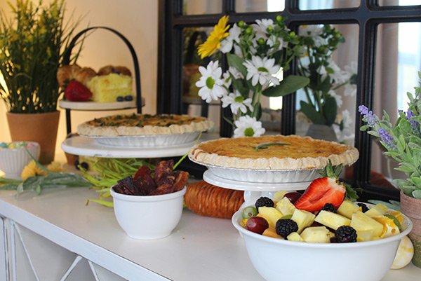 Breakfast spread on a white table with fruit, pies, and flowers, set near a window.