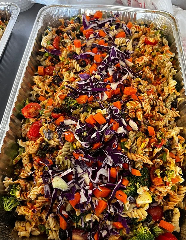 Pasta salad with rotini pasta, cherry tomatoes, chopped carrots, red cabbage, broccoli, and other vegetables in a large aluminum tray.