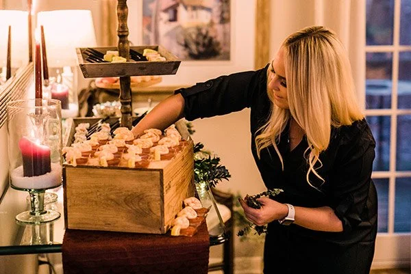 A woman arranging small desserts in a wooden box at a cozy indoor event.