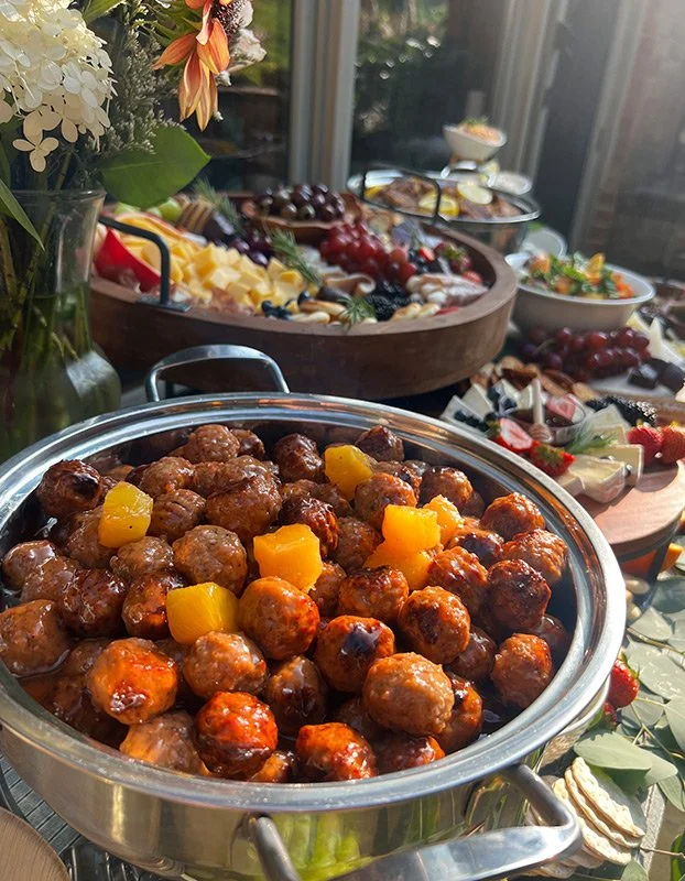 A stainless steel serving dish filled with glazed meatballs and pineapple chunks, set on a table with a spread of various appetizers, fruits, and cheese in the background, with sunlight streaming in.