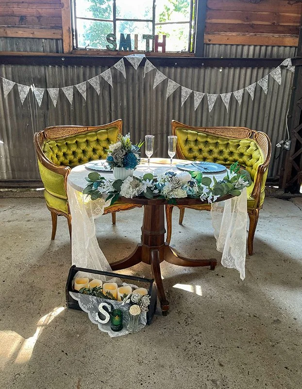 A wedding or special event table setup with a white lace tablecloth, floral centerpiece with white and blue flowers, two champagne flutes, and blue placemats. Two vintage green velvet upholstered chairs are behind the table. In front of the table, th