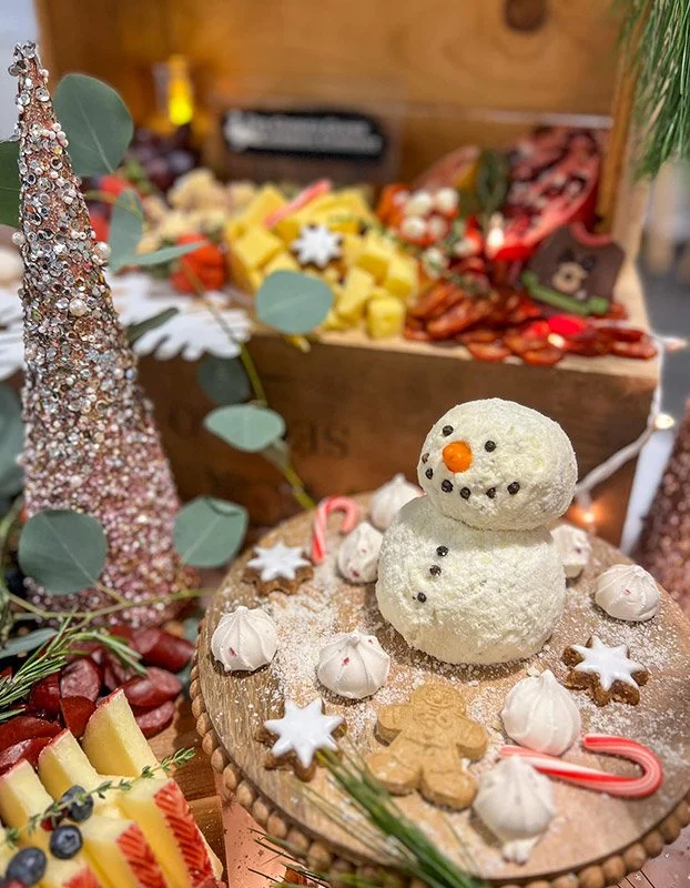 A festive holiday display with a snowman cake on a wooden round, surrounded by meringue cookies, candy canes, and gingerbread cookies. In the background, a crate holds colorful cheese and meat snack trays, with a decorative pink glittery Christmas tr