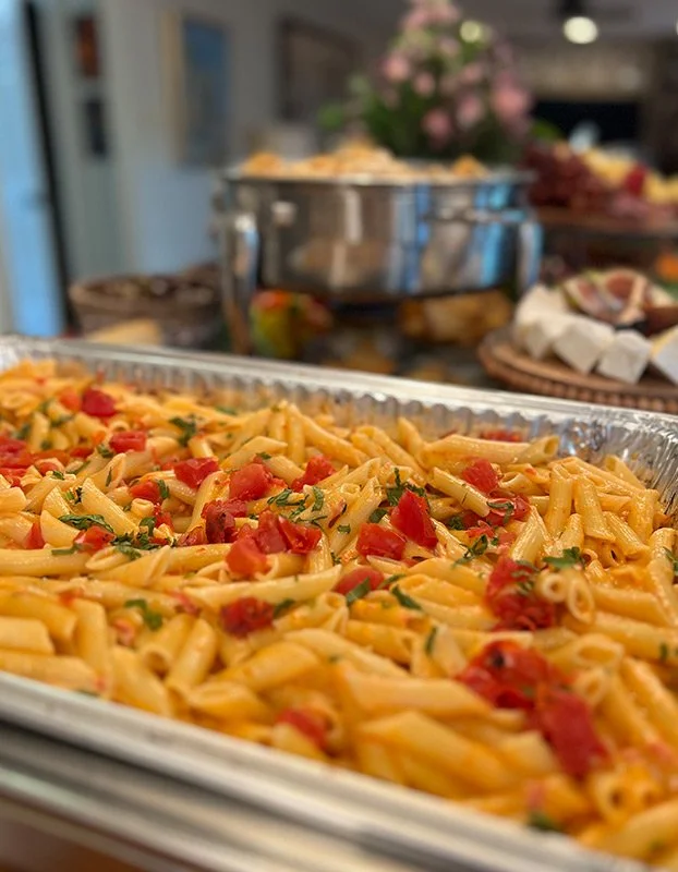Baked pasta with tomatoes and herbs in a metal tray, with a blurred background of a buffet table and a floral centerpiece.