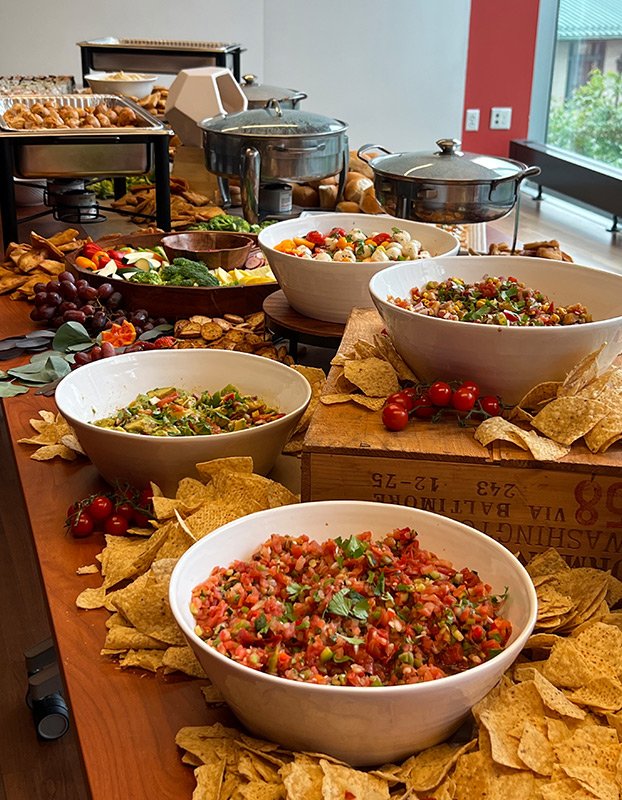 Buffet table with bowls of fresh salads, chips, and cherry tomatoes, along with serving pans in the background, set in a bright room with large windows.