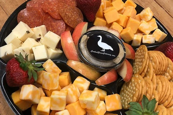 A cheese and charcuterie platter with assorted cheeses, sliced strawberries, apple slices, salami, crackers, cubed cheddar cheese, cubed Swiss cheese, and some cheese spread in a small container.