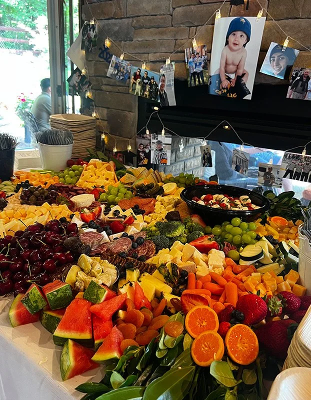 A fruit and cheese platter with grapes, watermelon, oranges, strawberries, and assorted cheeses, displayed on a table at a gathering, with photos hanging on a stone fireplace behind.