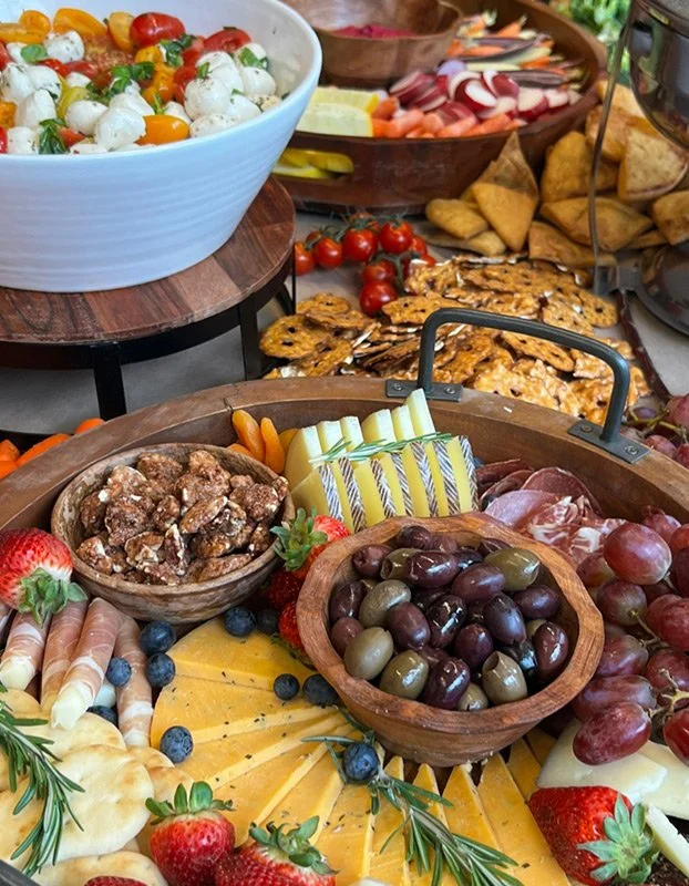 Assorted cheeses, grapes, strawberries, blueberries, nuts, and charcuterie on a table, with vegetable and dip platter and crackers in the background.