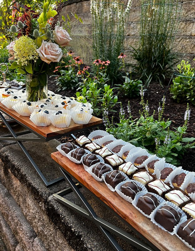 Tray of white cupcakes decorated with blueberries, and tray of chocolate-covered cookies, set on outdoor tables in a garden with flowers and greenery.