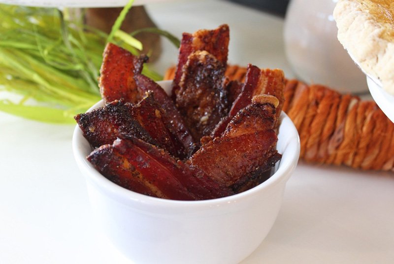 Bowl of cooked bacon strips on a white surface with greenery and bread in the background.