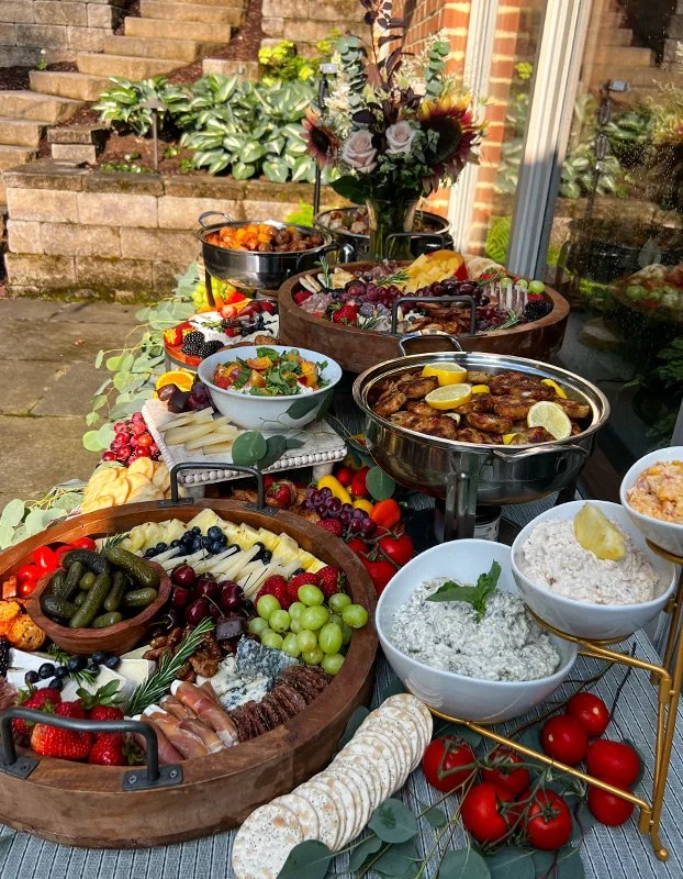 An outdoor table with a variety of foods including a cheese and charcuterie board, fresh grapes, cherry tomatoes, bowls of dips and spreads, grilled chicken, and a colorful fruit salad, set next to a glass window and a brick staircase.