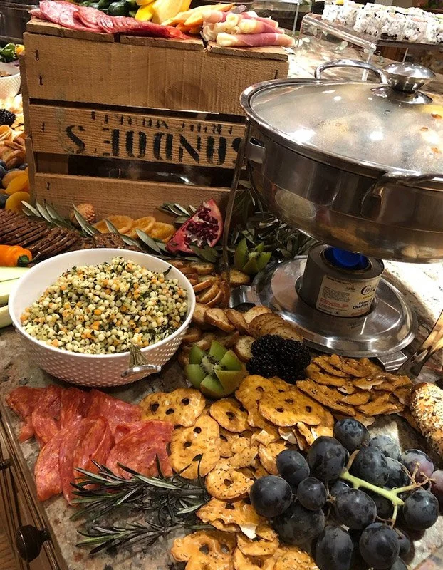 A buffet table with a variety of food, including fresh fruit, salad, cured meats, crackers, and a large chafing dish. There is also a wood crate in the background among the food display.