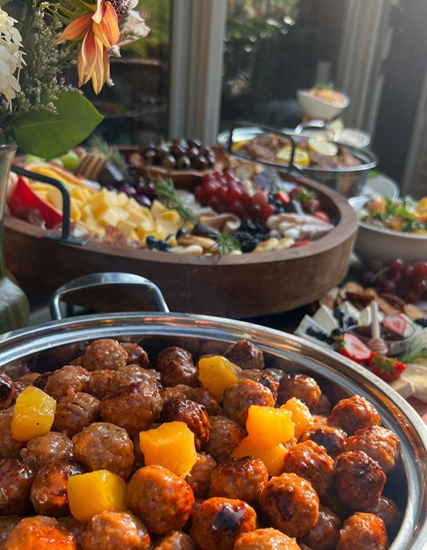 Close-up of glazed meatballs topped with chunks of pineapple on a serving dish. In the background, a large wooden platter holds cheese, grapes, and various appetizers, with bowls of salad and other dishes on a table near a window.