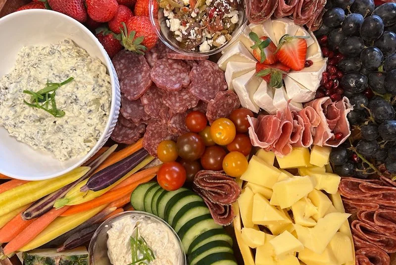 Assorted platter featuring cheeses, cured meats, cherry tomatoes, sliced cucumbers, colorful carrots, strawberries, grapes, and various dips.