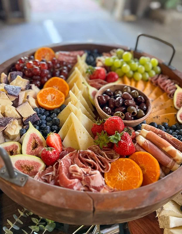 A large round wooden platter with a variety of fresh fruits, cheeses, cured meats, and olives arranged in sections, including strawberries, oranges, grapes, figs, blueberries, and black olives.