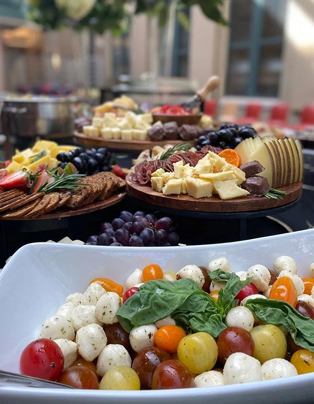A variety of cheese, grapes, and meat on platters, and a bowl of cherry tomatoes with basil leaves, likely part of a buffet or outdoor gathering.