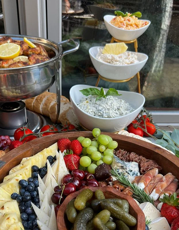 A spread of appetizer and snack foods, including fresh fruits like grapes, strawberries, cherries, and blueberries, various cheeses, cured meats, and dips, with bowls of spreads or salads and a roasted chicken with lemon slices on a table by a window