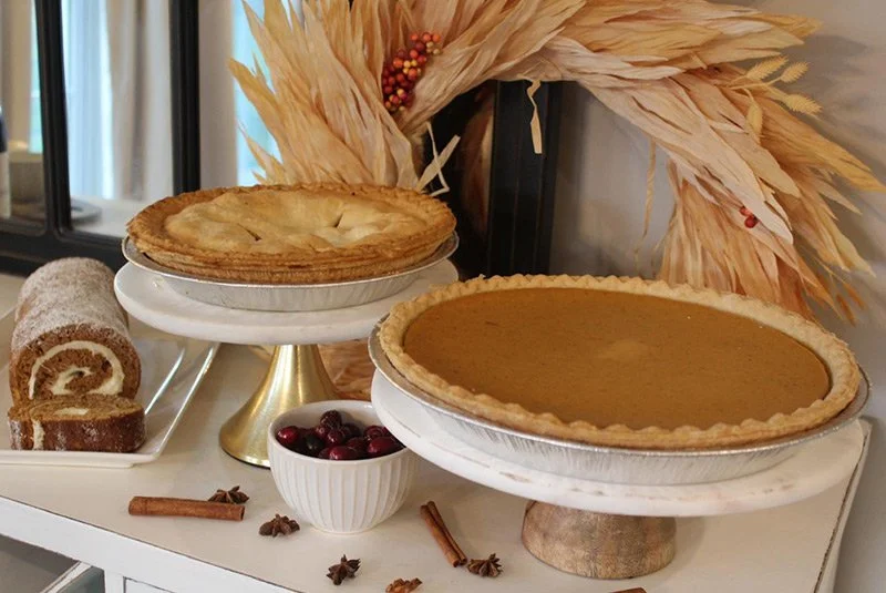 Two pies on display, one with apple filling and the other with pumpkin filling, along with a Swiss roll cake, a bowl of cranberries, cinnamon sticks, and star anise, decorated with dried pampas grass in the background.