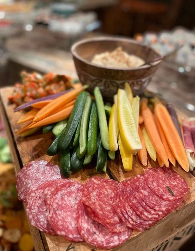 A wooden board of sliced salami, assorted fresh vegetable sticks including carrots, cucumbers, yellow bell peppers, and celery, with a bowl of hummus in the background.