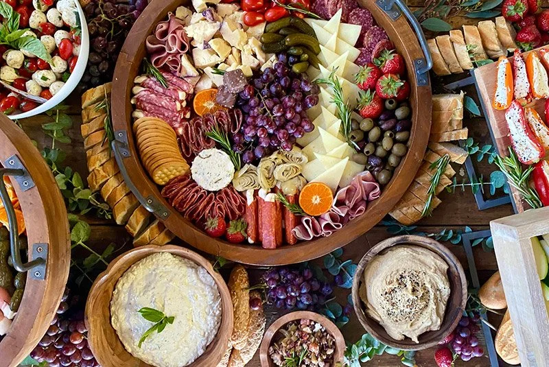 Cheese, charcuterie, grapes, strawberries, crackers, and sliced vegetables on a round wooden platter with additional dips and spreads surrounding it.