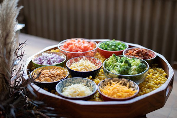 A wooden tray with small bowls of chopped vegetables and toppings, including diced tomatoes, chopped green onions, chopped red onions, shredded cheese, broccoli, corn, and other ingredients.