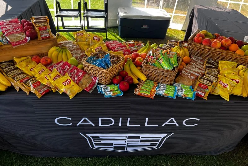 A table with an assortment of fresh fruits and snack packs, including bananas, apples, peaches, and granola bars, arranged on a black Cadillac-branded tablecloth at an outdoor event.