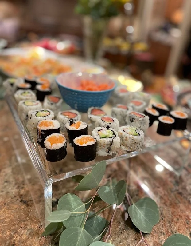 A display of assorted sushi rolls on a glass stand with a blue bowl of ginger in the background, set on a marble countertop with eucalyptus leaves underneath.