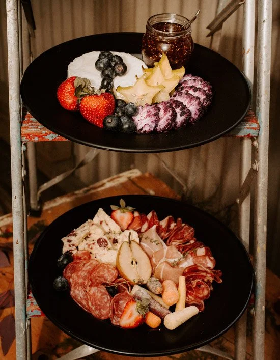 Two black plates with assorted breakfast foods on a metal rack, including fresh berries, cheese, sliced meats, and jam.