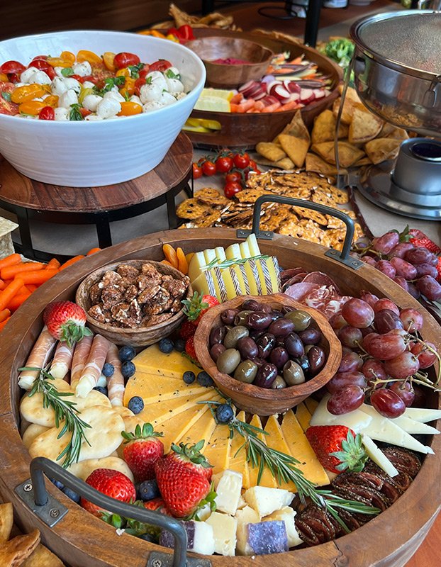 A variety of cheese, grapes, strawberries, olives, and cured meats arranged on a wooden charcuterie board, with a fruit salad bowl and other snacks in the background.