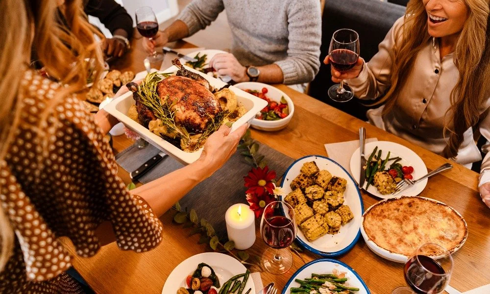 People gathered around a dinner table with a roast turkey, salads, corn casserole, pizza, and wine.
