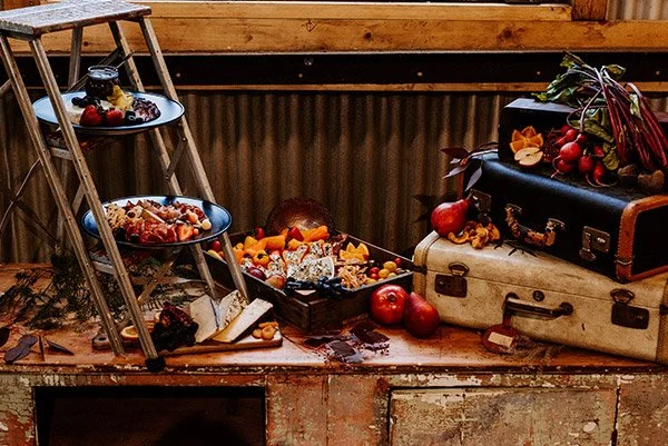 A rustic wooden table with vintage suitcases, fruit, and a black tray with various foods, including apples, berries, and cheese, decorated with flowers and greenery.