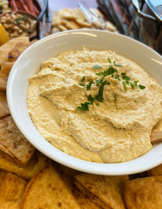 A white bowl filled with creamy hummus garnished with chopped parsley, surrounded by pita chips and snacks on a tray.