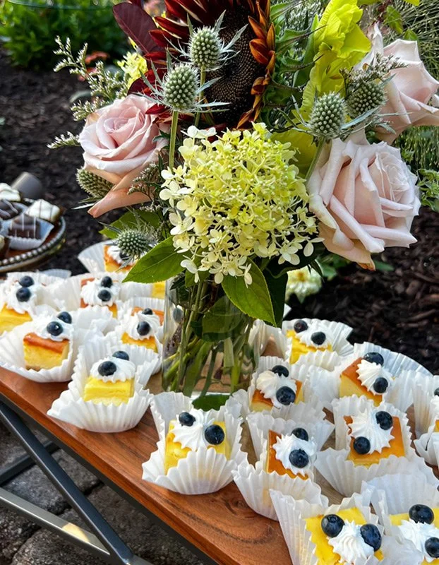 Vase with mixed flowers including roses, thistle, and hydrangeas on a wooden table surrounded by mini cheesecakes topped with blueberries and whipped cream.