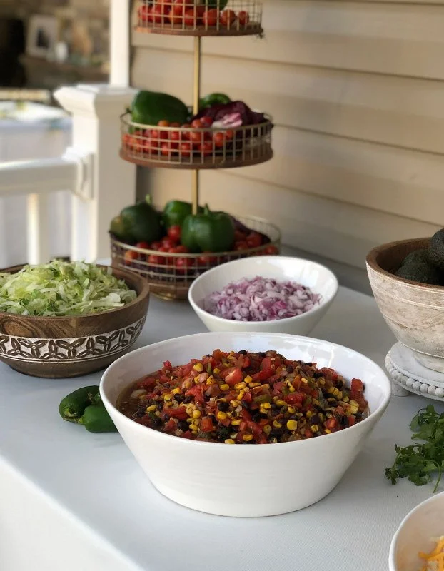 Bowl of chopped lettuce, bowl of diced red onions, bowl of chopped cilantro, bowl of salad with ground meat, tomatoes, corn, and black beans, and a tiered stand with bell peppers and other vegetables on a white tablecloth.