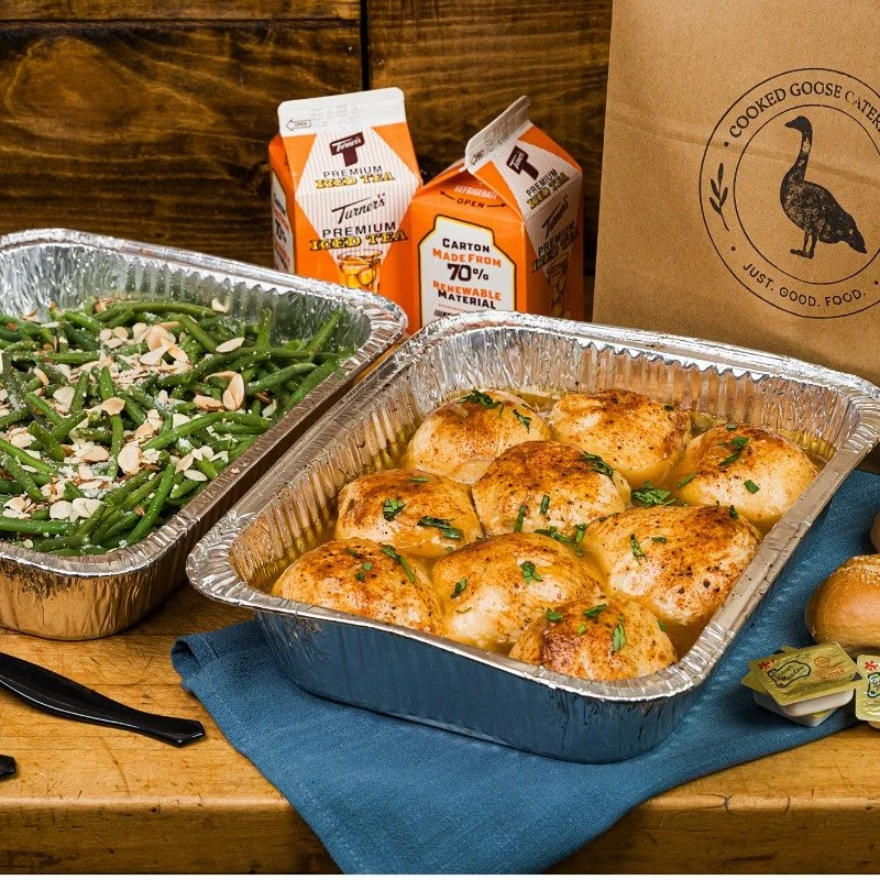 Aluminum tray of baked chicken with herbs in a sauce, a tray of green beans with sliced almonds, and a paper bag from Cooked Goose catering on a wooden table.