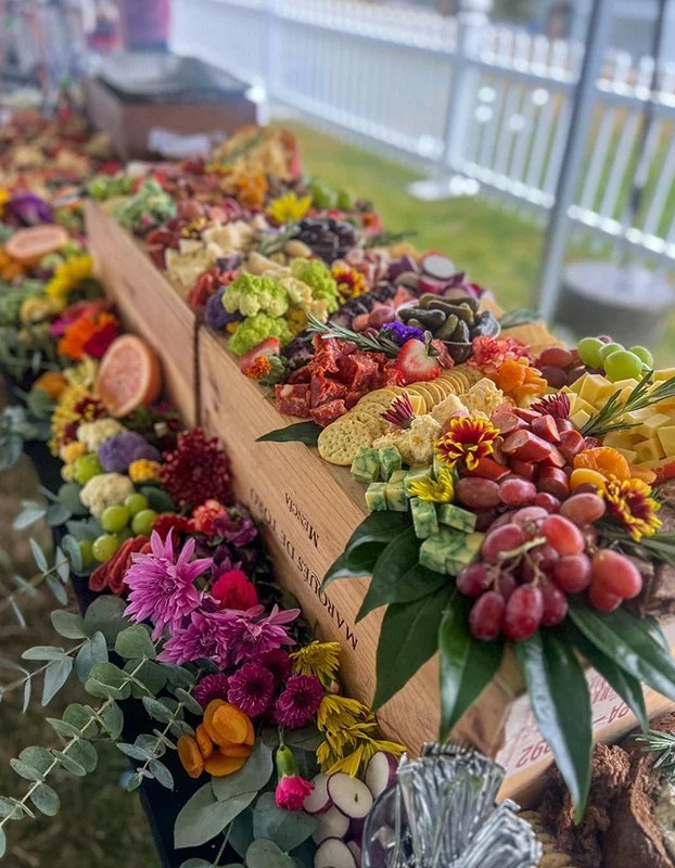 A long wooden table filled with colorful fruits, vegetables, and flowers, displayed on a porch or outdoor setting with a white railing and green grass in the background.