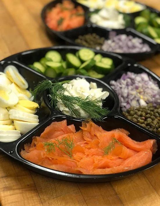 A black divided serving tray with various fresh vegetables and toppings, including smoked salmon garnished with dill, sliced cucumbers, hard-boiled eggs, capers, diced red onion, and a dollop of mayonnaise or sour cream, on a wooden surface.