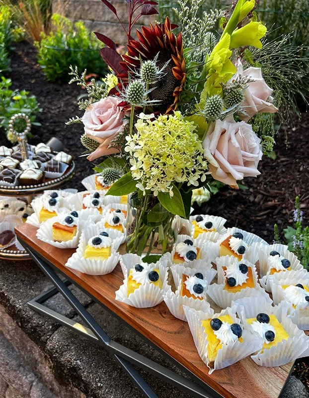 A floral arrangement with pink roses, yellow lilies, red and brown proteas, and various greens, placed on a wooden tray surrounded by cupcakes topped with blueberries and cream, set outdoors on a stone surface.