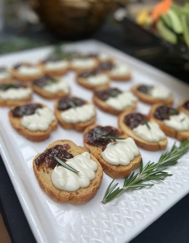 Slices of toasted baguette topped with creamy cheese, fig jam, and a sprig of rosemary on a white serving platter.
