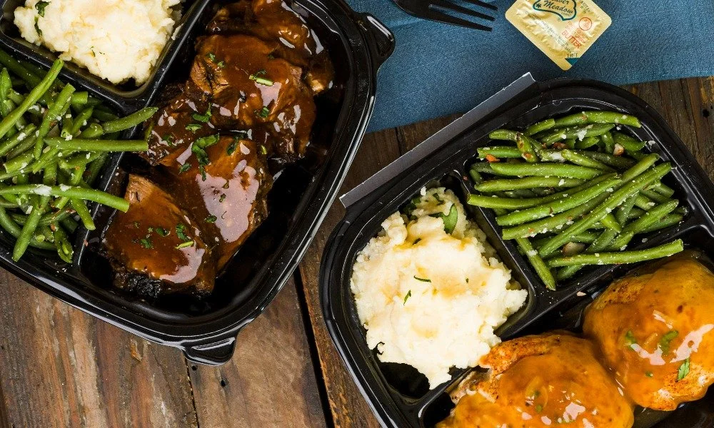 Two meal prep containers with mashed potatoes, green beans topped with grated cheese, and roast beef covered in gravy on a wooden table.